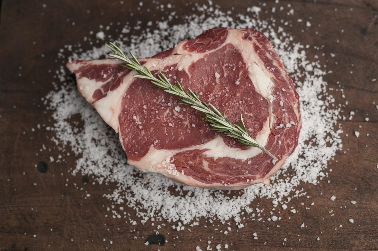 A raw ribeye with rosemary on a dark board scattered with coarse salt